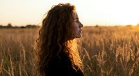 Young woman with curly hair in profile enjoying golden hour sunset in wheat field, peaceful moment in nature for wellness and mindfulness concepts.