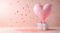 Pink heart balloon floating above silver gift box with bow on pink background with golden stars and glitter, romantic festive celebration setup.