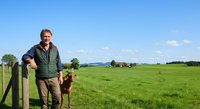 Mature male farmer standing by wooden fence with brown dog in expansive green pasture under clear sky, suitable for agriculture and rural lifestyle content.