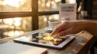 Person's hand touching digital tablet screen with glowing financial icons, coffee cup and notebook on wooden desk by window with warm sunlight, representing fintech, banking, investment and digital commerce.