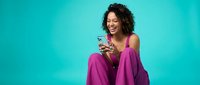 Happy young African American woman in pink outfit using smartphone while sitting against turquoise background, expressing joy and connection through technology.