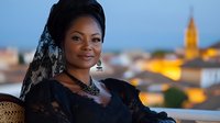 Young African woman wearing traditional black lace dress and headscarf smiles warmly at sunset, with blurred Mediterranean cityscape and church tower in background.