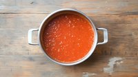 Fresh homemade tomato soup in metal cooking pot on rustic wooden table background, bright red color with visible seasoning and herbs floating on surface.