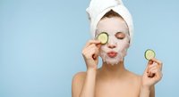 Young woman with white towel on head applying facial mask holding cucumber slices over eyes for skincare routine and spa treatment at home.