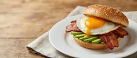 Gourmet breakfast burger with fried egg, crispy bacon and avocado slices on sesame bun served on white plate over rustic wooden table background.