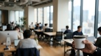 Young professional holding white coffee cup in modern office workspace with colleagues working at desks and city skyline visible through windows.