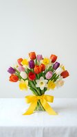 Colorful spring flower bouquet with tulips and daffodils in glass vase with yellow ribbon bow on white table for Mother's Day celebration and gifts.