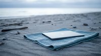 Abandoned blue fabric and white notebook lying on sandy beach with ocean and cloudy sky in background, suitable for loss, forgotten memories, travel writing, and editorial concepts.