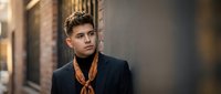 Young professional businessman in dark suit with orange scarf standing confidently on urban street. Corporate executive portrait for business marketing materials.