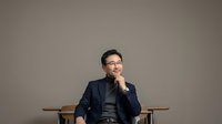 Thoughtful male professional wearing glasses and dark blazer sitting on wooden chair against neutral background, hand near chin in contemplative pose.