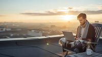Young businessman working on laptop computer on rooftop terrace during golden hour sunset with city skyline background for remote work and productivity.