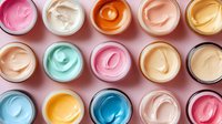 Colorful array of whipped cosmetic creams in jars viewed from above, featuring pastel pink, blue, mint, and yellow tones arranged in neat rows on pink background.