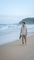 Caucasian male in casual linen shirt walking barefoot along sandy beach at sunrise, holding shoes, with ocean waves and coastal cliffs in background for lifestyle and travel content.