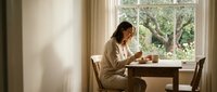 Young woman in white robe writing in journal at wooden table by window with natural light streaming in, peaceful morning routine and self-reflection moment.