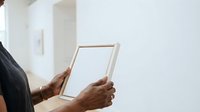 Person of African descent, male, wearing dark business attire, holding empty white frame against neutral wall background for interior design or home decor display purposes.