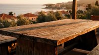 Wooden table surface with coastal Mediterranean landscape view, golden hour sunlight, rustic outdoor dining setting with sea and hillside architecture backdrop.