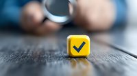 Yellow cube with checkmark symbol on wooden surface with blurred background showing magnifying glass, representing approval, verification and quality control.
