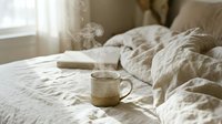 Cozy morning bedroom scene with steaming coffee mug on unmade bed, soft natural light streaming through window creating peaceful atmosphere for relaxation.
