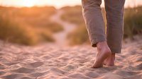 Barefoot person walking on sandy beach path at golden sunset, casual linen pants, warm summer evening, freedom and relaxation concept.