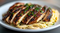 Grilled blackened chicken breast sliced and served over creamy fettuccine pasta garnished with fresh herbs, close-up view showing juicy meat texture and seasoning.