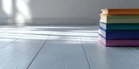 Stack of colorful hardcover books on light wooden floor with soft window shadows, minimalist education and reading concept background.