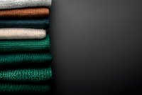 Stack of folded knitted sweaters in various colors including green, orange, beige and teal arranged on dark background with copy space for seasonal fashion concepts.