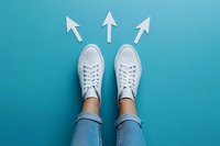 White sneakers with upward pointing arrows on turquoise background, wearing blue jeans. Top view of feet symbolizing direction, choice, and forward movement.