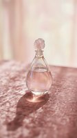 Elegant glass perfume bottle with rose quartz crystal stopper filled with pink liquid, displayed on textured mauve surface with soft natural lighting for fragrance and beauty product marketing.