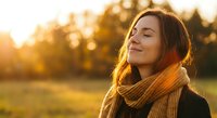 Young woman with closed eyes enjoying warm sunlight in autumn park wearing cozy knit scarf during golden hour sunset creating peaceful mindful moment outdoors.