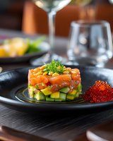 Fresh salmon tartare layered with diced avocado and mango, garnished with microgreens and red caviar, served on black plate in upscale restaurant setting.
