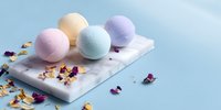 Colorful bath bombs on marble tray with scattered dried flower petals against blue background, perfect for spa and self-care products.