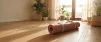 Rolled yoga mat on wooden floor in bright living room with plants and natural sunlight streaming through windows for home fitness and wellness practice.