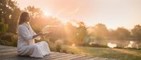 Young woman in white robe meditating peacefully on wooden deck during golden hour sunrise, practicing mindfulness and wellness in serene natural setting.