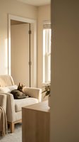 Cozy modern living room interior with tabby cat relaxing on beige armchair near window with natural light streaming through blinds.