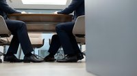 Two businessmen in dark suits sit across from each other at a wooden table during a corporate meeting or negotiation discussion in a modern office environment.