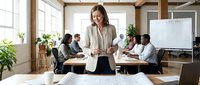 Middle-aged Caucasian female business professional leading corporate training session with diverse team in modern office environment with plants and whiteboard.