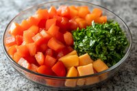 Fresh diced red bell peppers, cubed mango and chopped parsley in glass bowl on granite counter, ready for cooking or salad preparation.