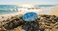 Blue gemstone resting on rocky surface at tropical beach with turquoise ocean waves and golden sand creating serene coastal meditation scene.