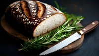 Fresh artisanal bread with sesame seeds sliced on wooden board, served with rosemary sprigs and bread knife against dark background, moody food photography.