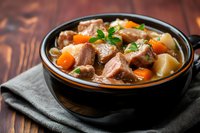 Hearty beef stew with tender meat chunks, carrots and potatoes in rich brown gravy, garnished with fresh parsley, served in rustic black bowl on wooden table.