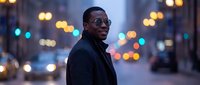 Confident young man with dark skin wearing sunglasses and dark jacket stands on urban street at dusk with blurred city lights in background.