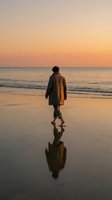 Person in brown coat walking toward ocean at sunset, reflected in wet sand beach. Solitude and contemplation concept.