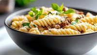 Fusilli pasta with ground meat sauce and parmesan cheese garnished with fresh basil leaves in black ceramic bowl, close up view on light background.