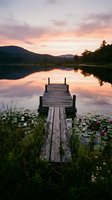 Wooden dock extending into calm lake at sunset with mountain reflections and pink sky creating peaceful lakeside scene for relaxation and nature themes.