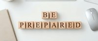 Wooden letter blocks spelling "BE PREPARED" arranged on white desk with notebook and computer mouse for business planning and readiness concepts.