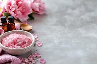 Pink sea salt crystals in ceramic bowl with peony flowers and essential oil bottles for spa relaxation and aromatherapy wellness treatments.