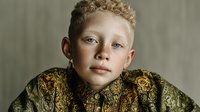 Young blonde child with curly hair wearing ornate golden embroidered jacket against neutral background for fashion and portrait photography.