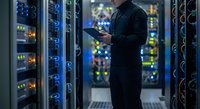 IT technician monitoring server performance on tablet in modern data center with blue LED lighting and network equipment racks for cloud computing operations.