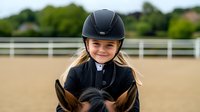 Young female equestrian in black helmet and riding outfit smiling while sitting on horseback in sandy arena with white fence and green trees in background.
