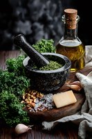 Fresh kale pesto ingredients with black mortar, olive oil, parmesan, nuts and garlic on rustic wooden board against dark background. Food preparation scene.